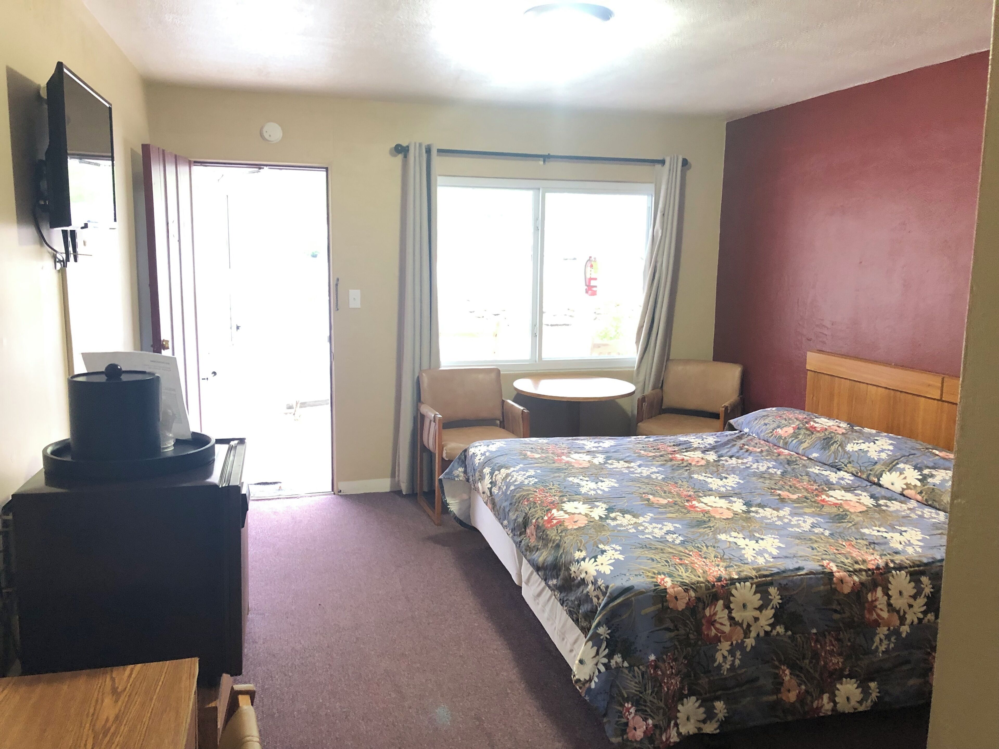 A hotel room with a bed, a desk, a chair, a small table, a television, and a window with curtains.