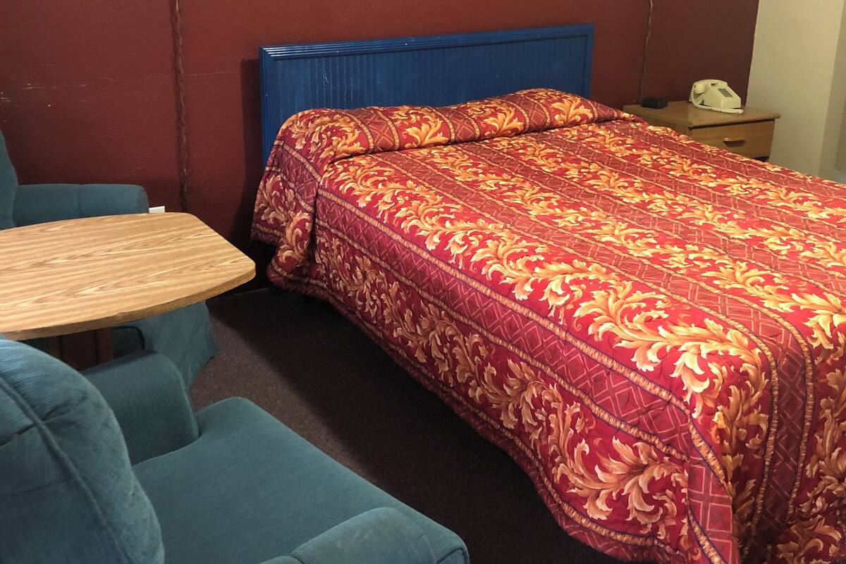 Une pièce comprenant un lit, un fauteuil bleu, une table en bois et deux lampes fixées au mur.