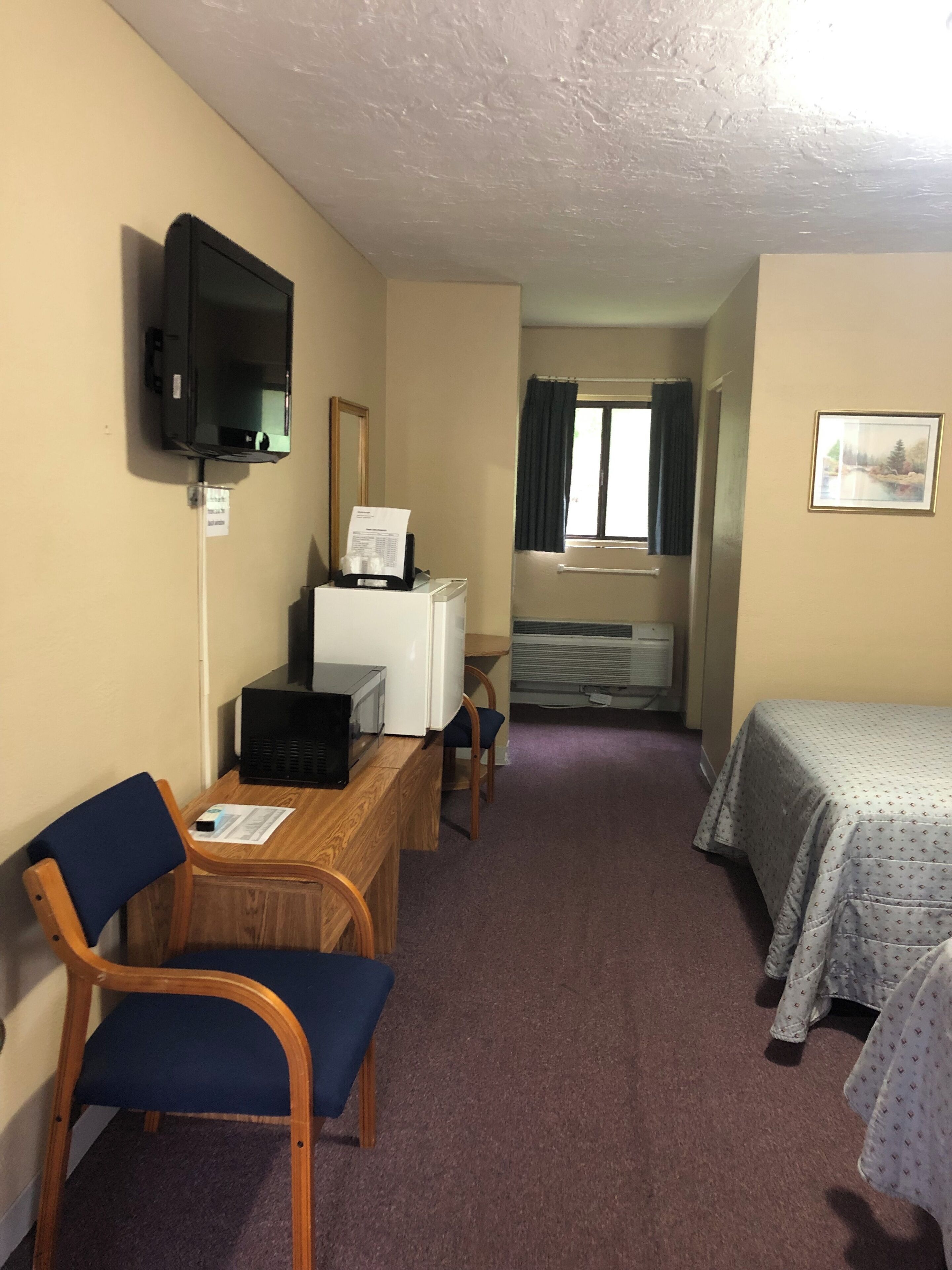A hotel room with a wooden desk, a chair, a bed with a patterned bedspread, a television mounted on the wall, and a window with curtains.
