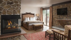 A bedroom with a stone fireplace, a bed with a headboard, a bench, a TV mounted on a stone wall, and a view through a glass door. - The Landing at Estes Park - Riverside Retreat (Estes Park)