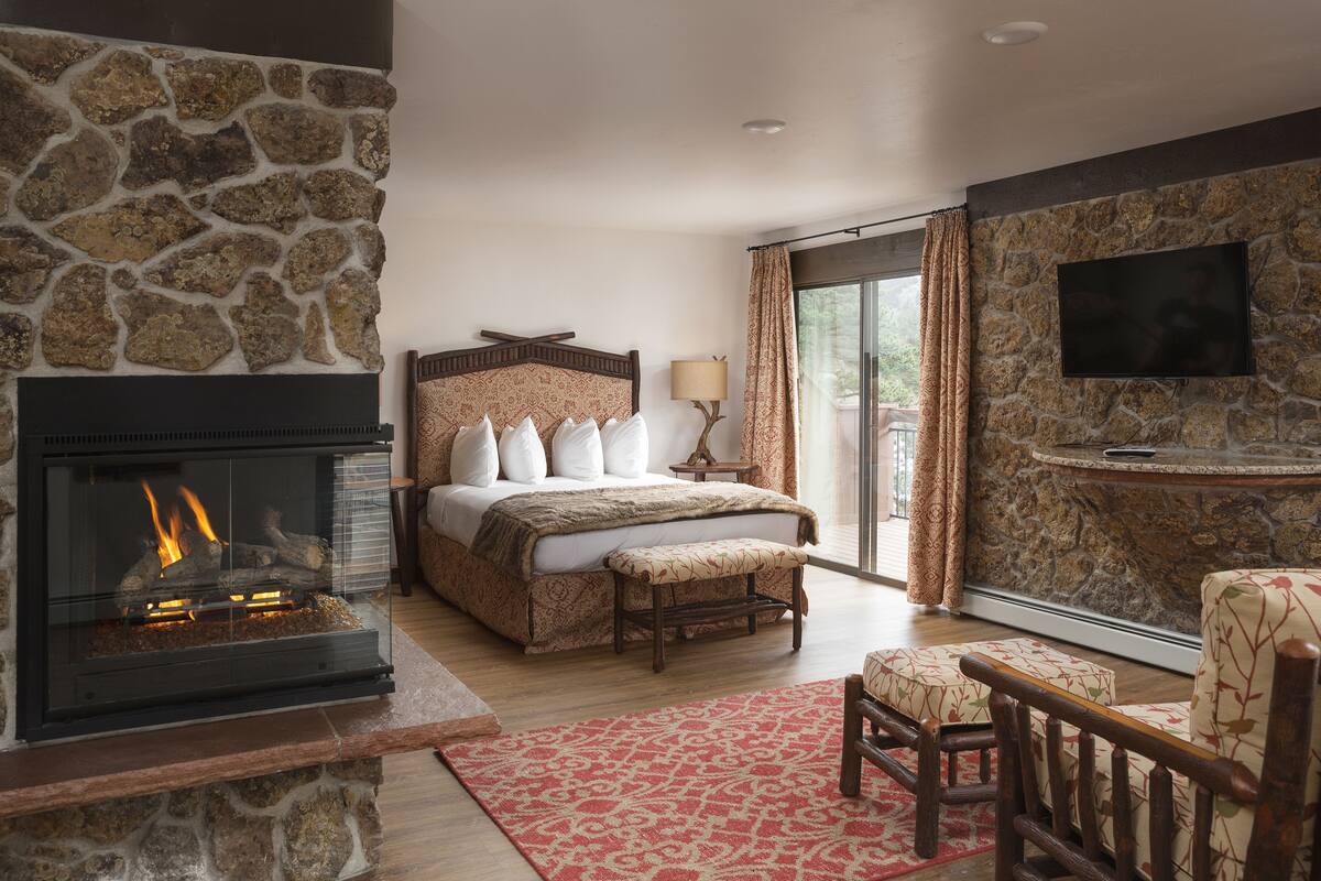 A bedroom with a stone fireplace, a bed with a headboard, a bench, a TV mounted on a stone wall, and a view through a glass door.