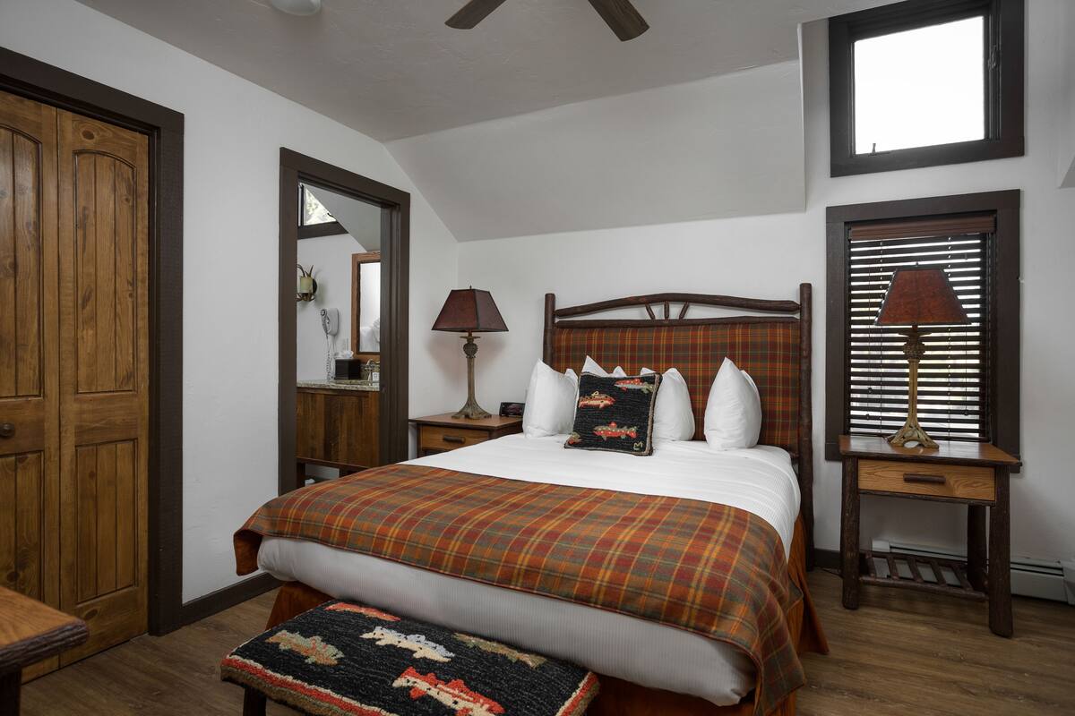 A bedroom with a large bed, wooden headboard, two bedside tables with lamps, a wooden door, and a window with blinds.