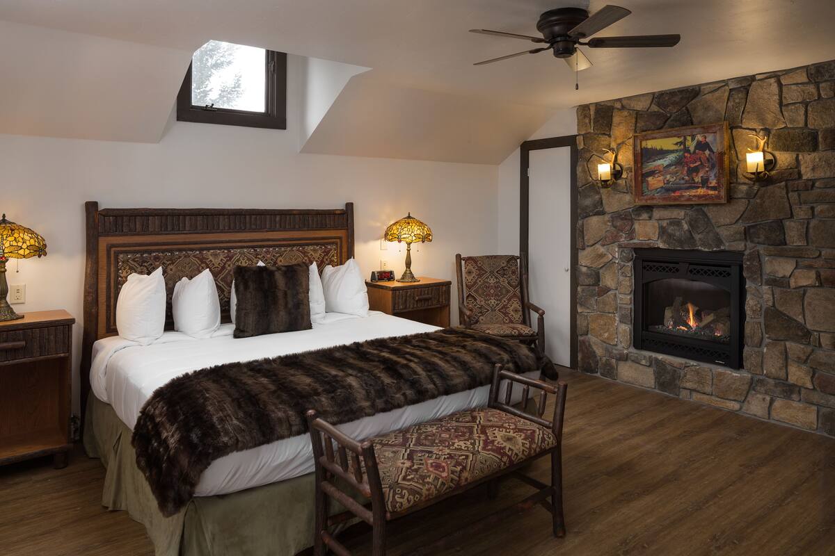 A bedroom with a large bed, a stone fireplace, a ceiling fan, and a nightstand with a lamp.