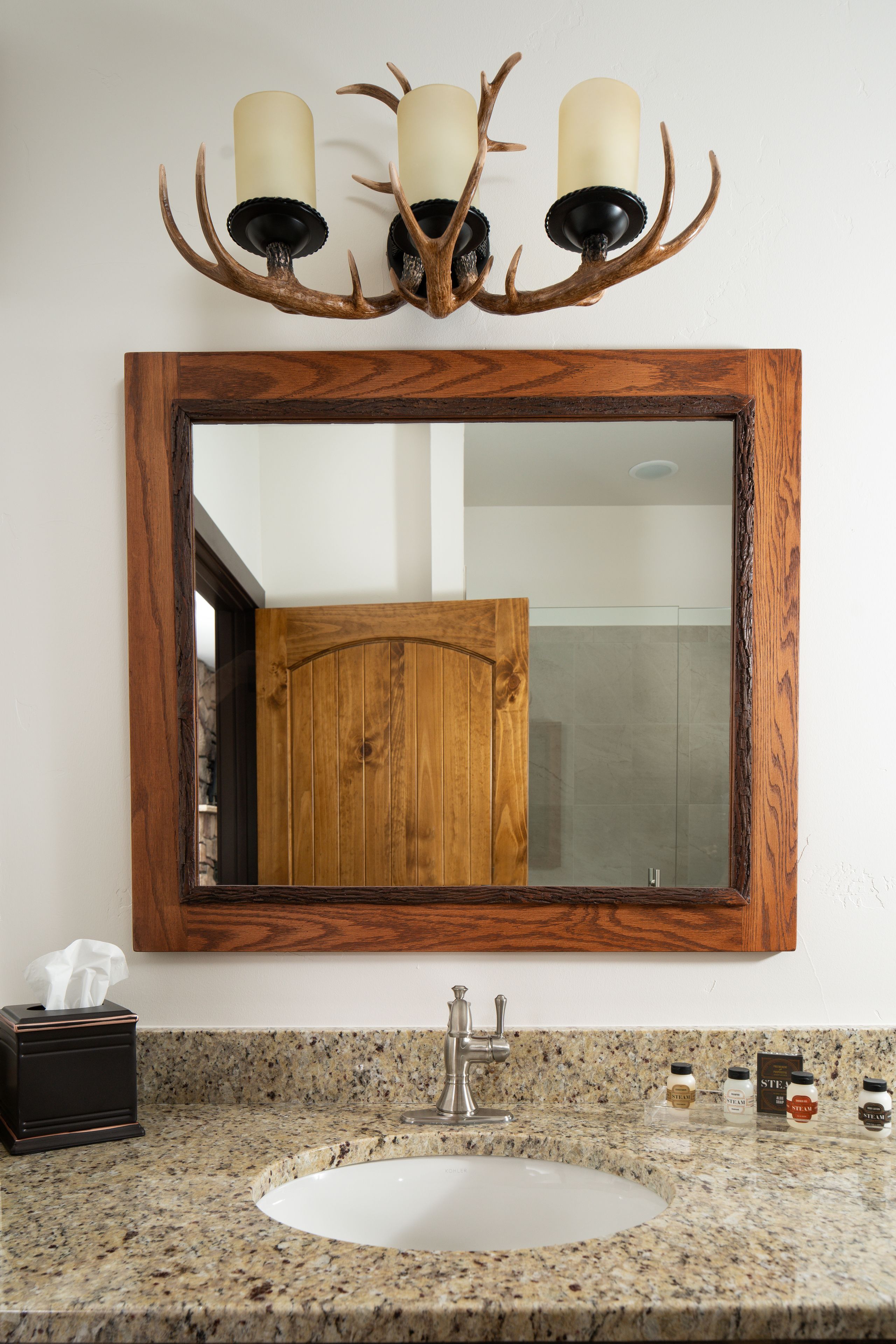 A bathroom with a wooden-framed mirror, a granite countertop, a sink, a tissue box, and a deer antler chandelier.
