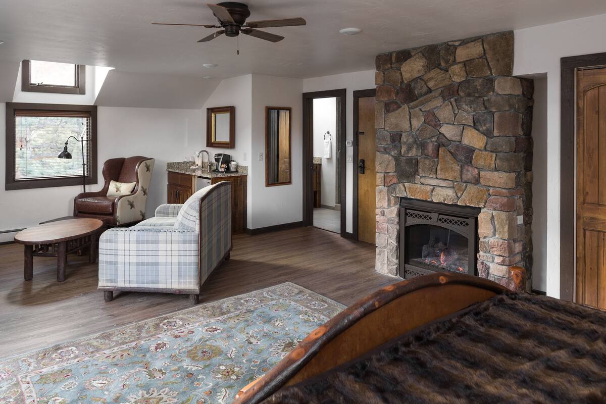 A living room with a stone fireplace, a ceiling fan, a plaid sofa, a wooden coffee table, and a patterned rug.