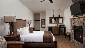 A bedroom with a large bed, a wooden nightstand, a lamp, a stone fireplace, a TV, and a kitchen area with a microwave and sink. - The Landing at Estes Park - Riverside Retreat (Estes Park)
