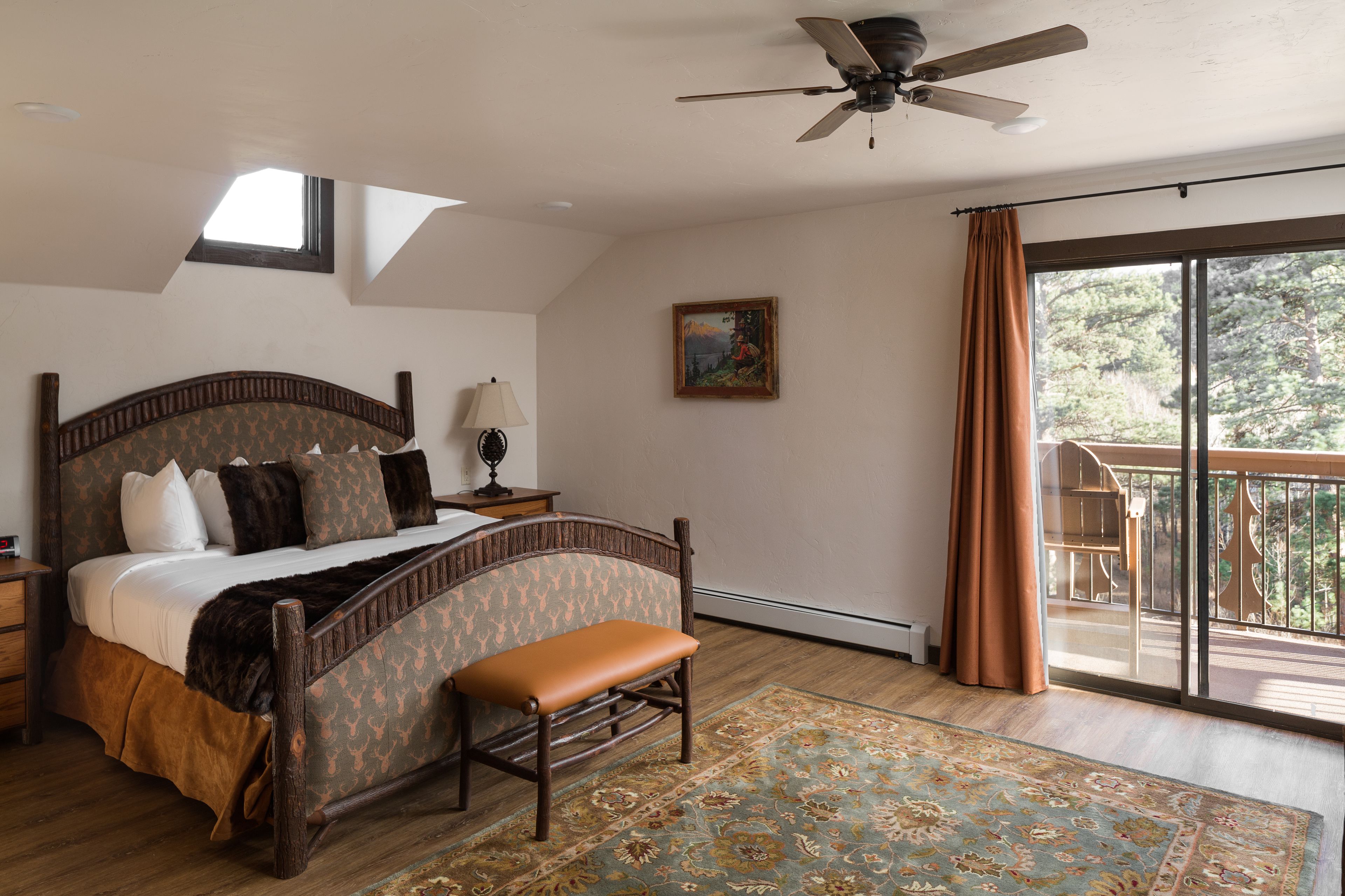 Une chambre avec un grand lit, un banc, un ventilateur de plafond, une table de chevet, une lampe, un tableau et un balcon avec des rideaux.