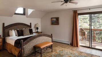 Une chambre avec un grand lit, un banc, un ventilateur de plafond, une table de chevet, une lampe, un tableau et un balcon avec des rideaux.