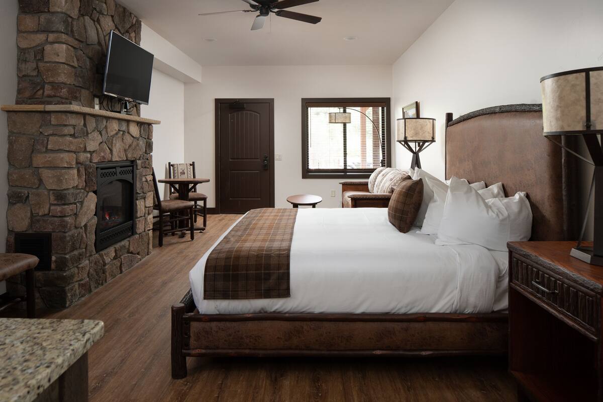 A bedroom with a stone fireplace, a bed with a headboard, a television mounted on the fireplace, a window with blinds, and a nightstand with a lamp.