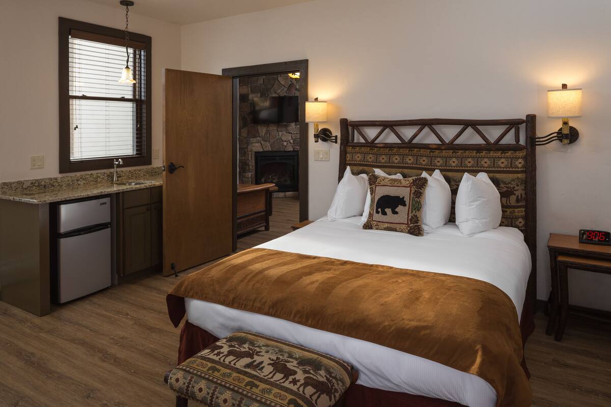 A bedroom with a bed, a nightstand, a lamp, a clock, a fireplace, and a small kitchenette.