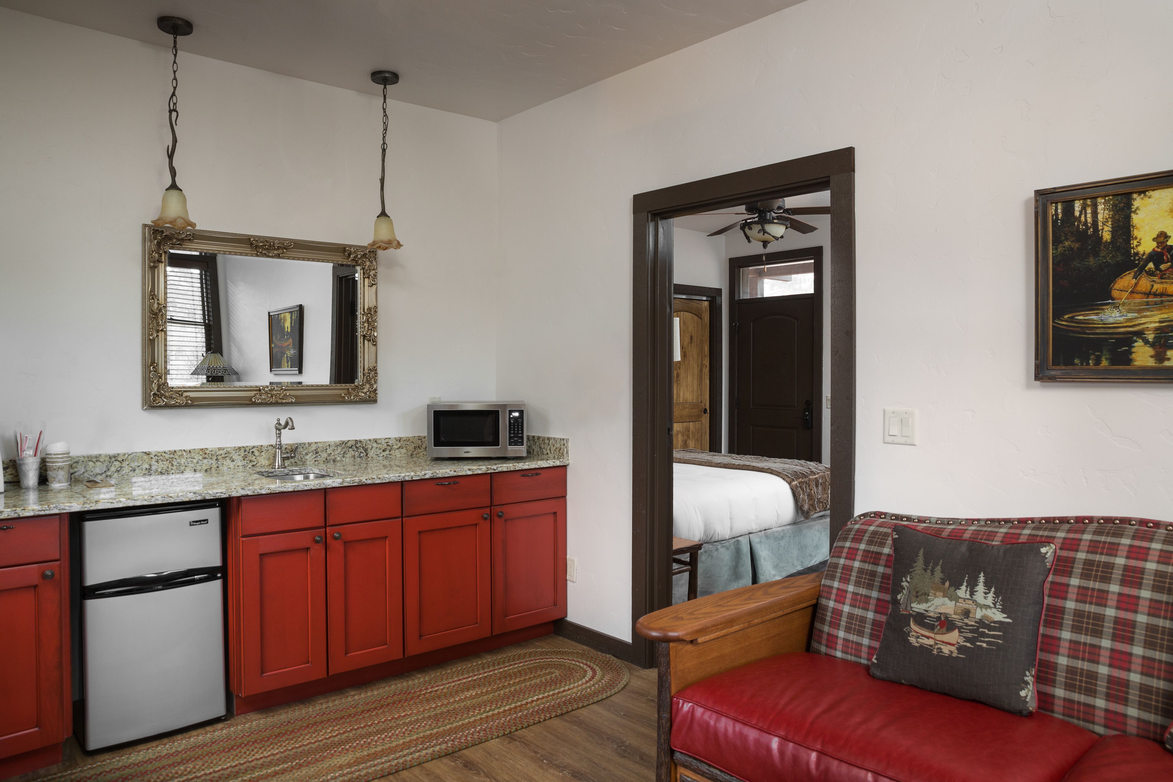 A compact kitchen with red cabinets, a microwave, and a refrigerator. There is a wooden dining table and a red couch with a decorative pillow.