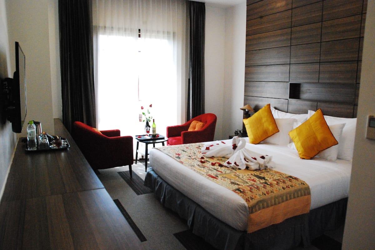 Une chambre d’hôtel avec un grand lit, deux fauteuils rouges, une petite table avec une bouteille et des verres, et un téléviseur fixé au mur.