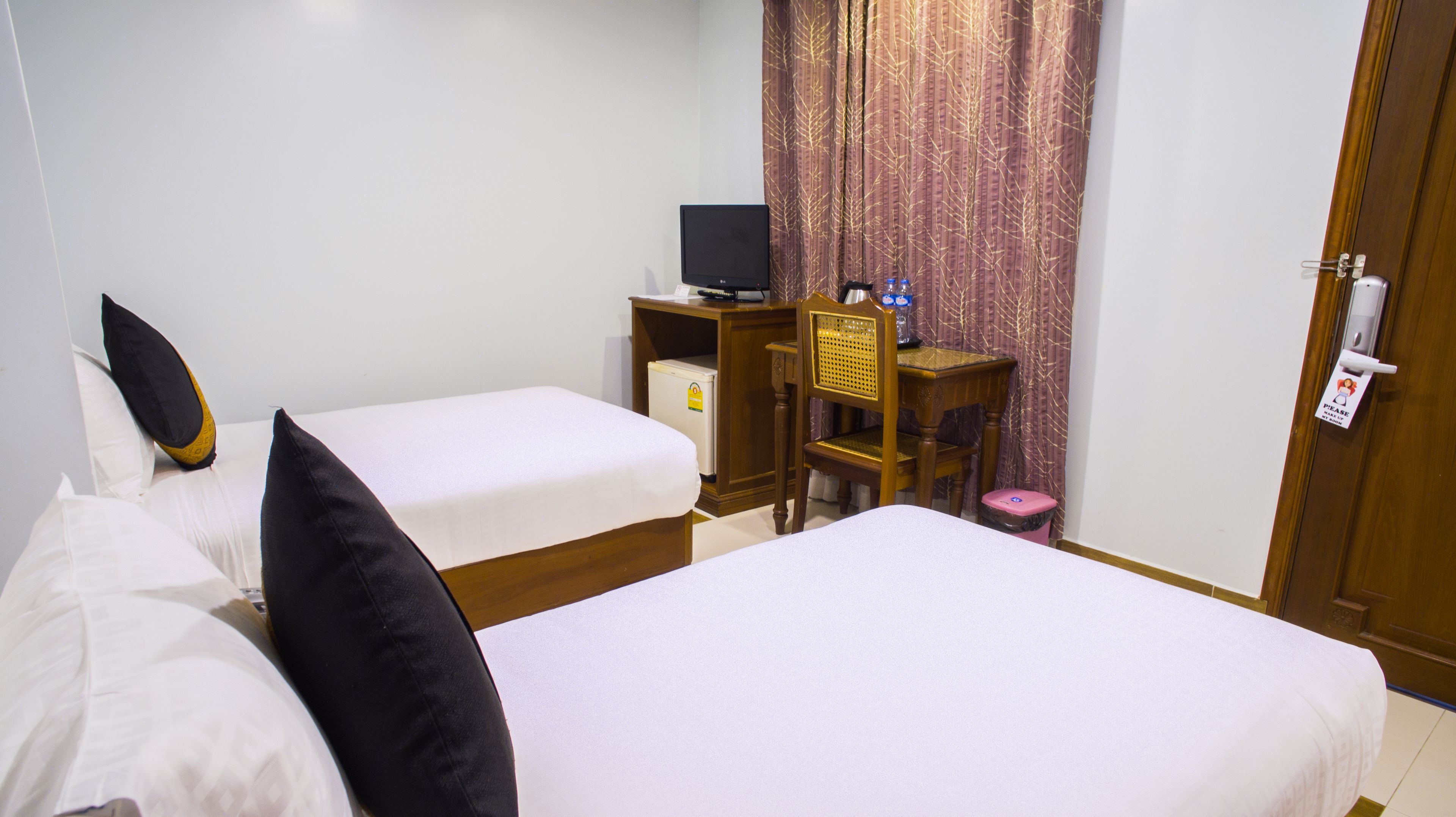 Habitación de hotel con dos camas individuales, un escritorio con computadora, un refrigerador pequeño y un cubo de basura.
