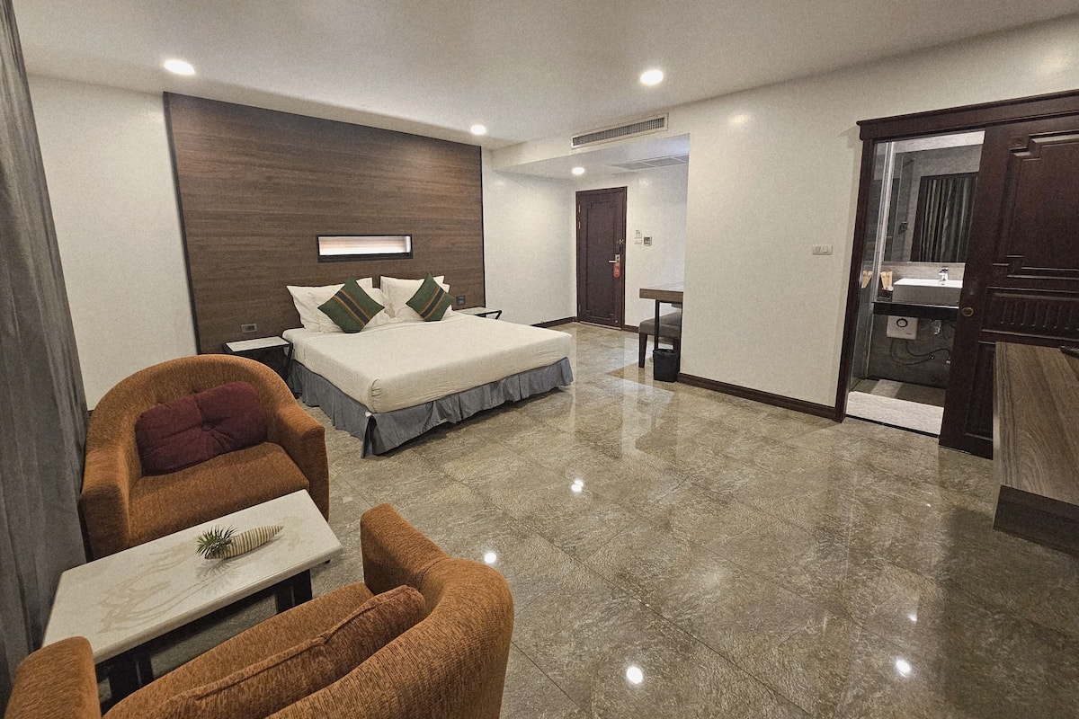 Une chambre d’hôtel moderne comprenant un lit, un coin salon avec une chaise et une table, une salle de bain avec un lavabo et un miroir, et une porte donnant sur une autre pièce.