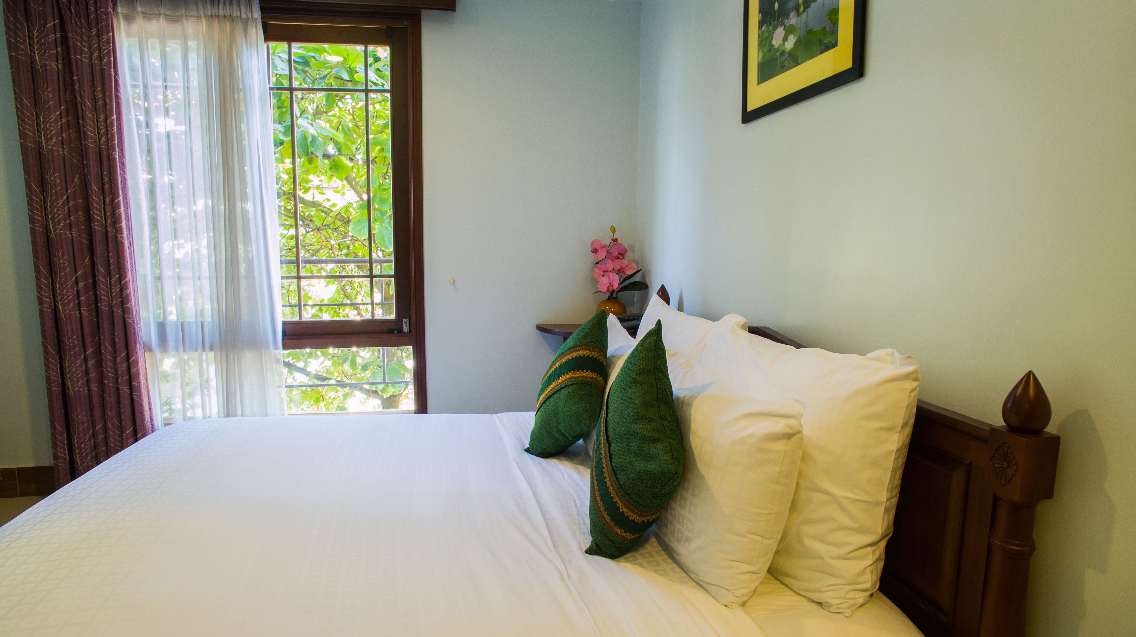 A neatly made bed with white linens and two green pillows, a window with a view of trees, and a framed picture on the wall.