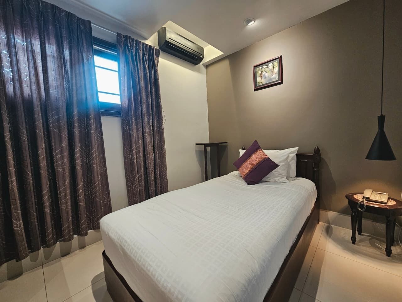 Habitación de hotel con cama, cortinas, equipo de aire acondicionado, mesita de noche con lámpara y un cuadro en la pared.