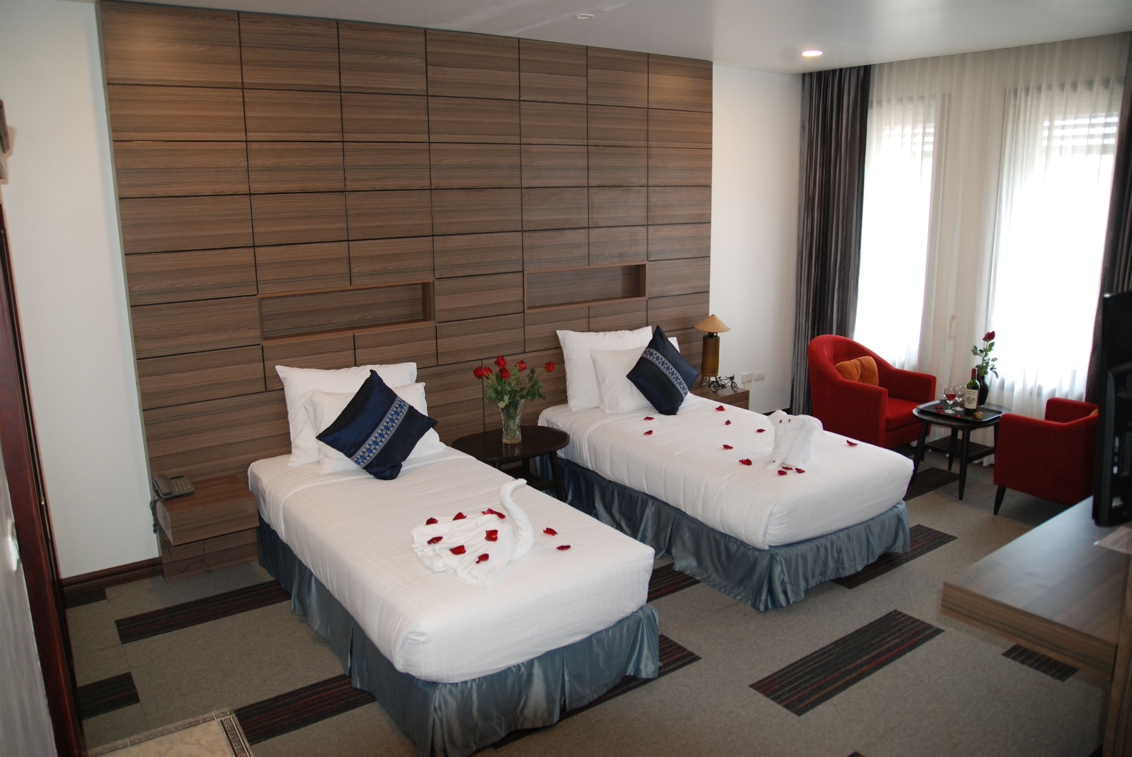 A hotel room with two beds, a wooden headboard, a red armchair, a small table with a lamp, and a view of the outdoors through the window.