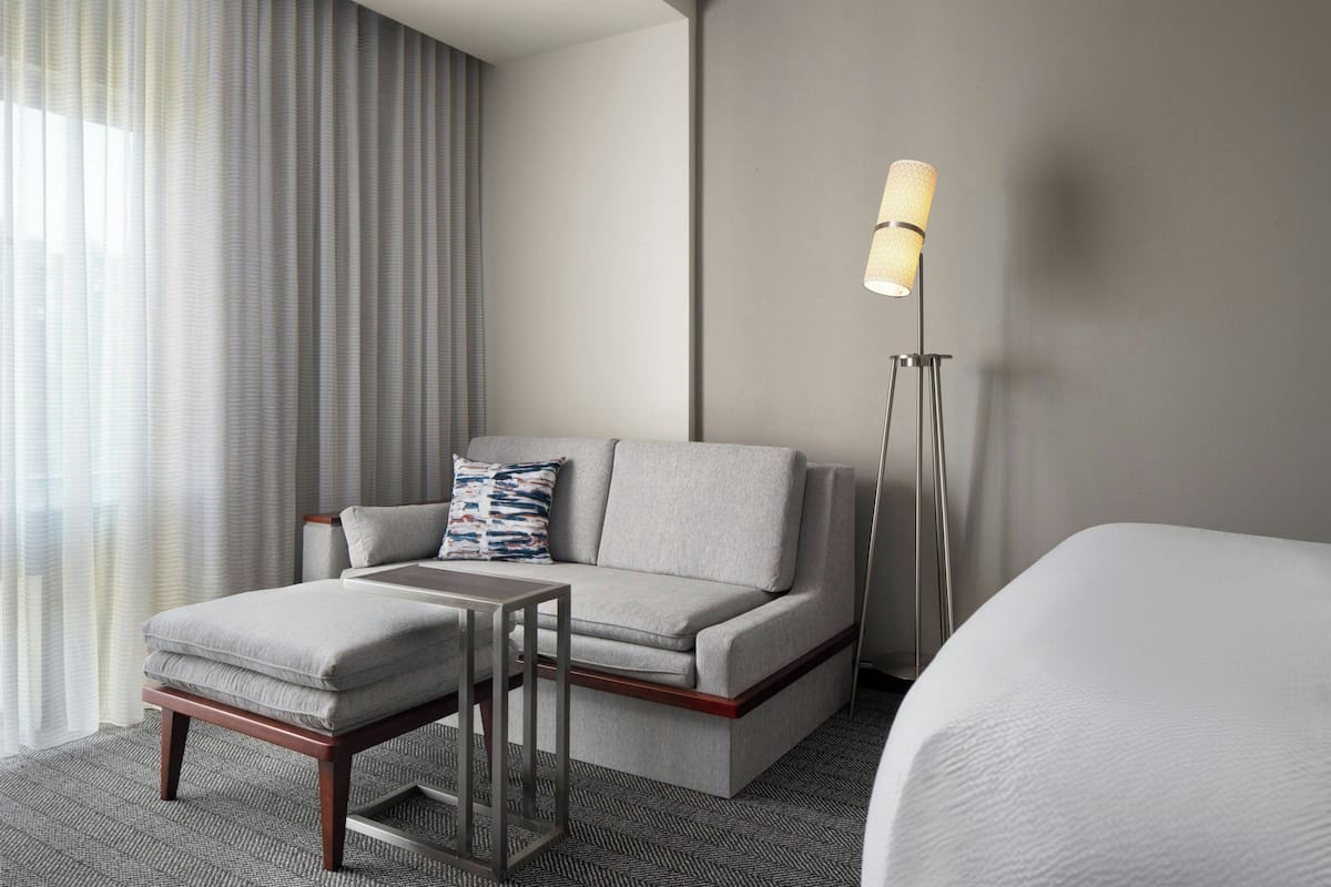 A modern hotel room with a sofa, a small table, a lamp, and a bed.
