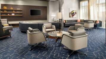 A modern hotel lobby with a large sectional sofa, multiple armchairs, a coffee table, and a flat-screen TV mounted on the wall.