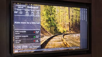 A TV screen displaying a weather forecast and stock market indices for Courtyard GoBoard, with a scenic forest view on the right.