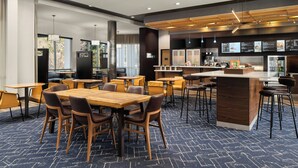 A modern hotel lobby with wooden tables and chairs, a central bar area, and a large window providing a view of the outdoors.