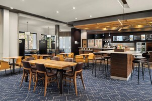 A modern hotel lobby with wooden tables and chairs, a central bar area, and a large window providing a view of the outdoors.