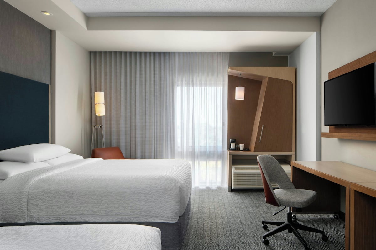 A hotel room with two beds, a desk, a chair, a TV, and a large window with curtains.