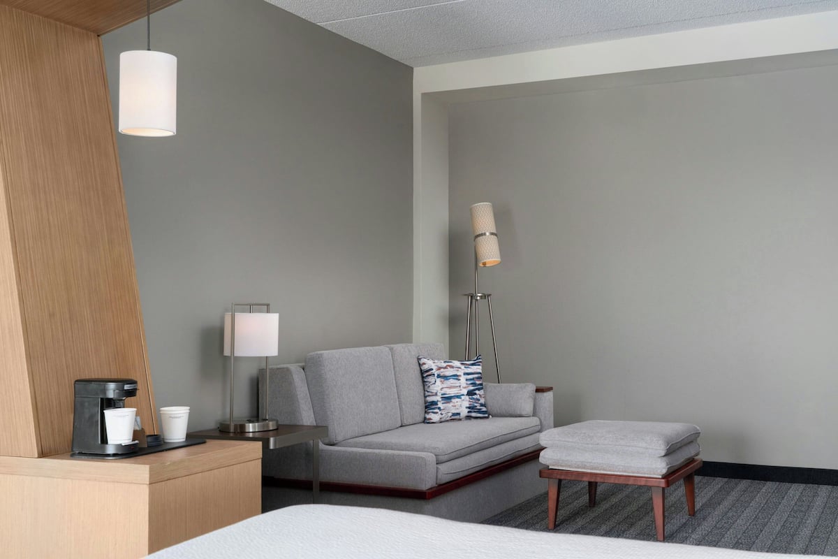 A hotel room with a bed, a grey sofa, a small table with a coffee maker, a lamp, and a floor lamp.