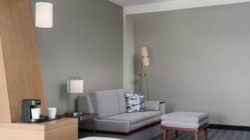 A hotel room with a bed, a grey sofa, a small table with a coffee maker, a lamp, and a floor lamp.