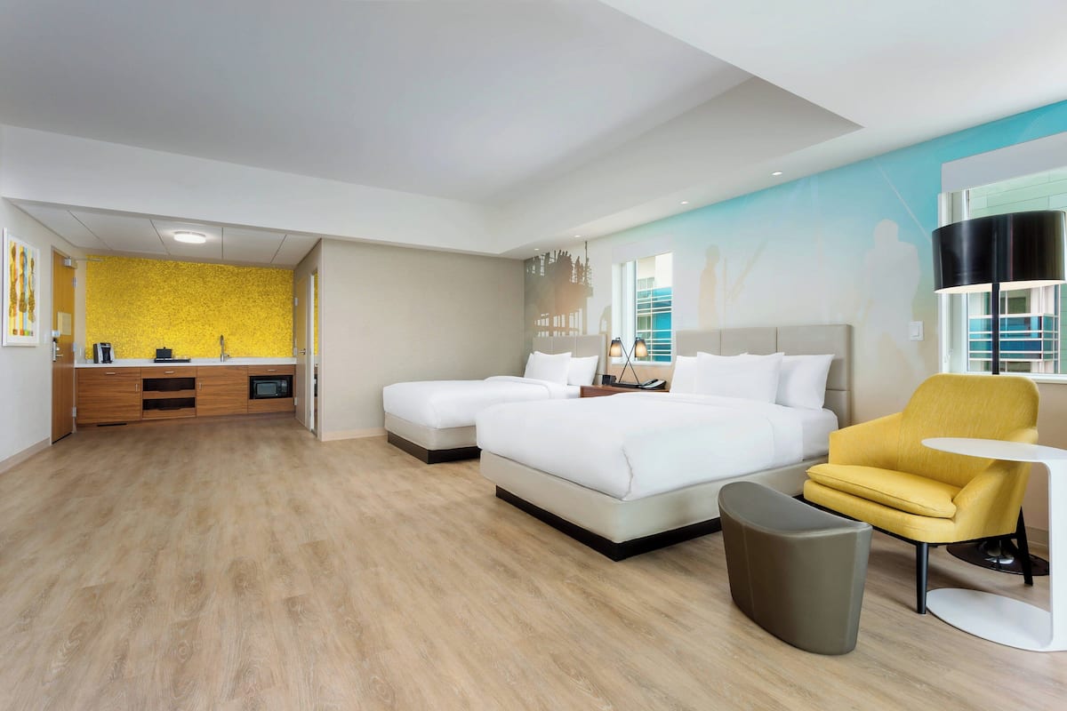 A modern hotel room with a wooden floor, two beds, a yellow armchair, a small table, and a large window with a view.