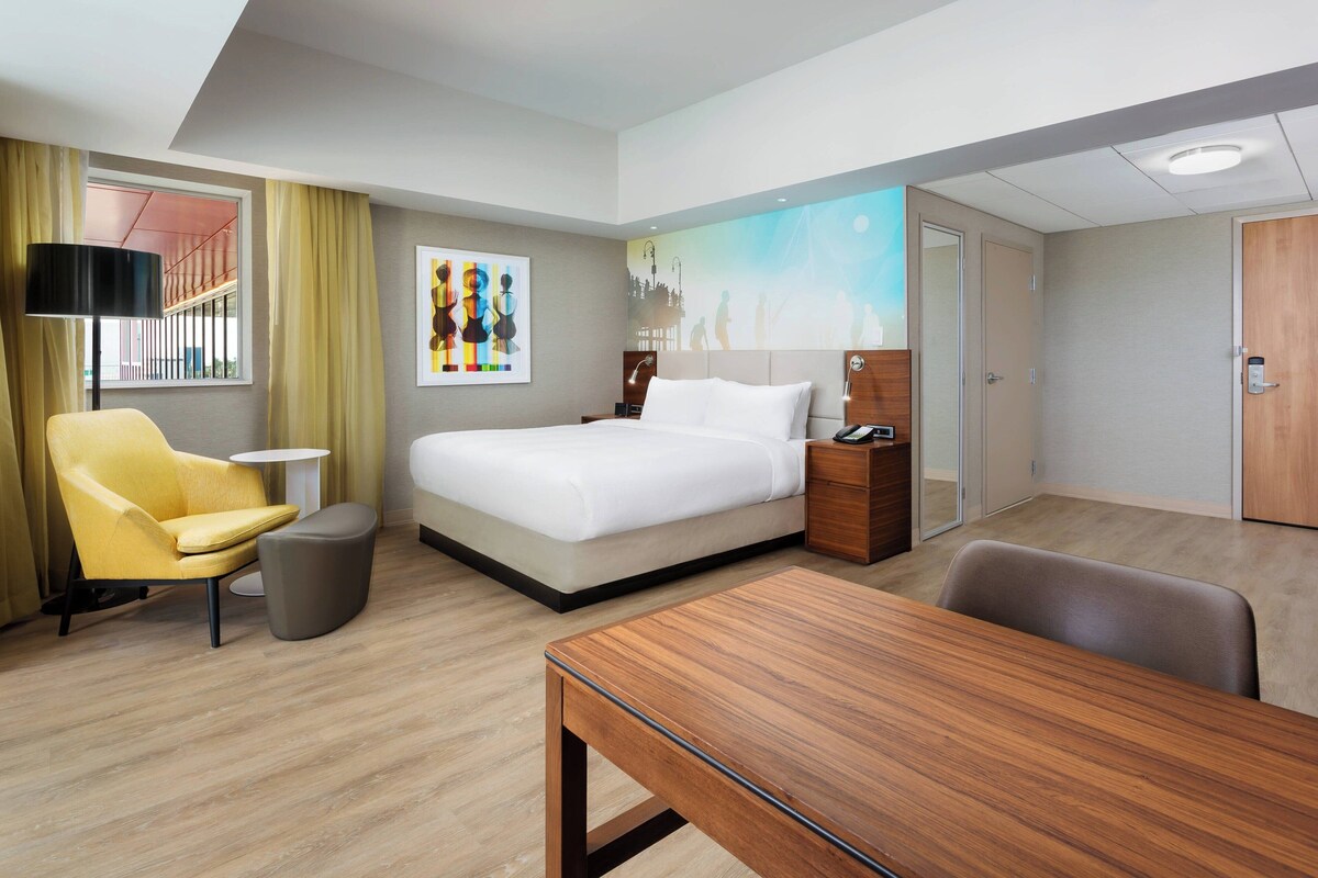 A hotel room with a bed, a desk, a chair, a lamp, and a painting on the wall.