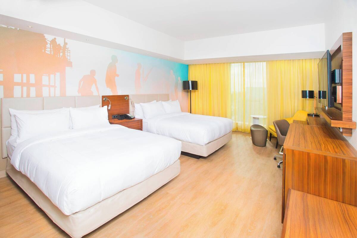 A hotel room with two beds, a wooden desk, a mirror, and a large window with yellow curtains.