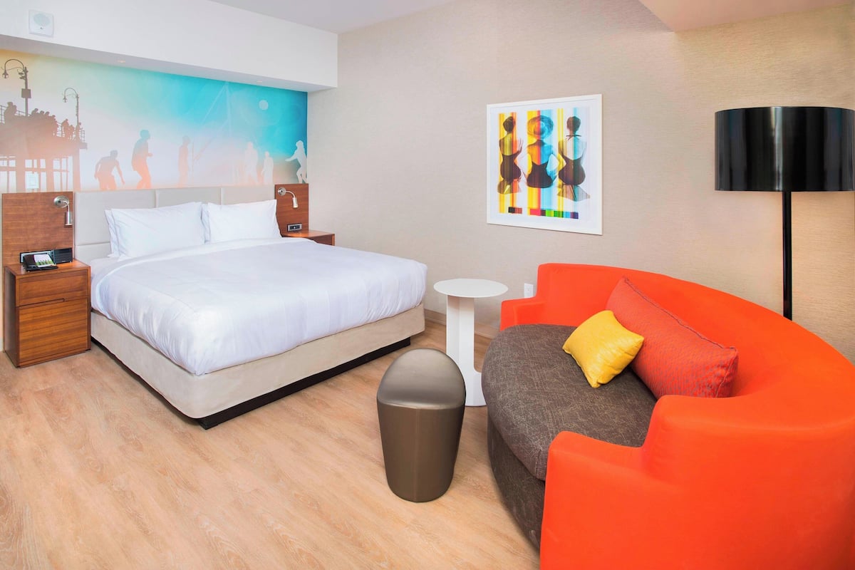 A modern hotel room with a large bed, a small round table, a sofa, and a trash bin.