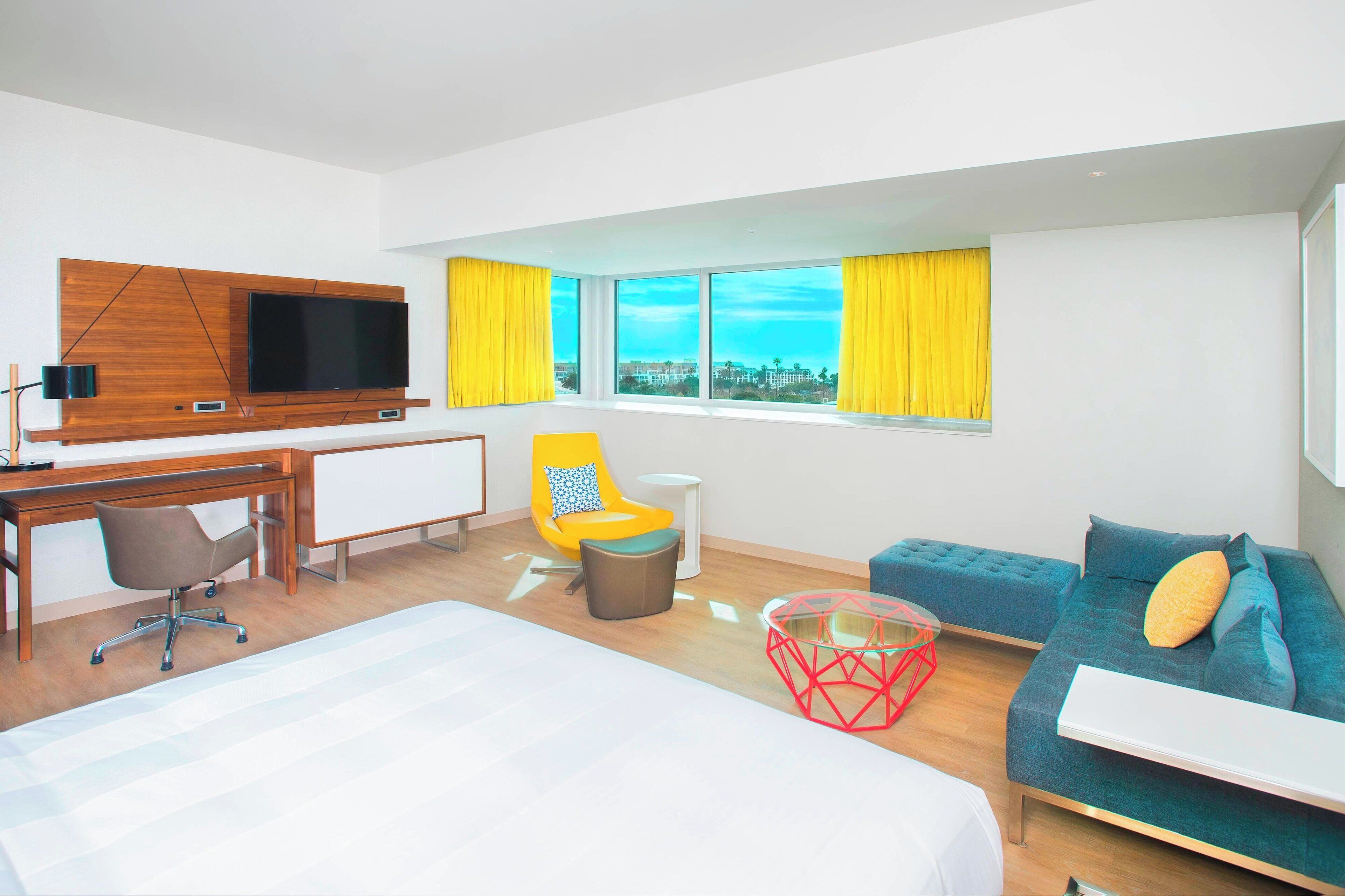 A modern hotel room with a large bed, a desk, a TV, a sofa, and a window with yellow curtains.