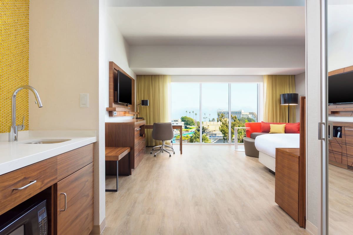 A hotel room with a kitchen area, a desk, a chair, a bed, and a large window offering a city view.