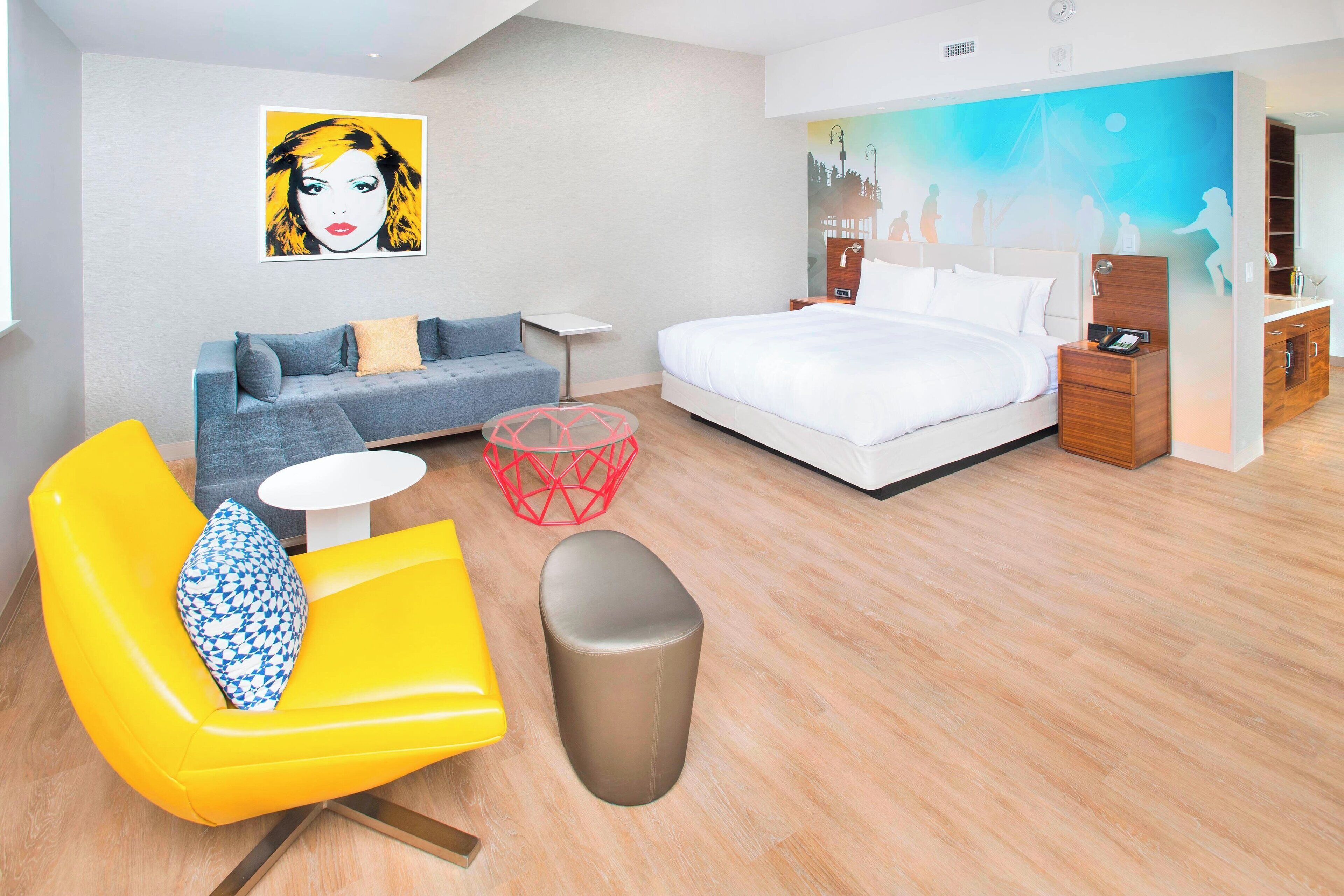 A modern hotel room with a bed, a sofa, a small round table, and a yellow armchair.