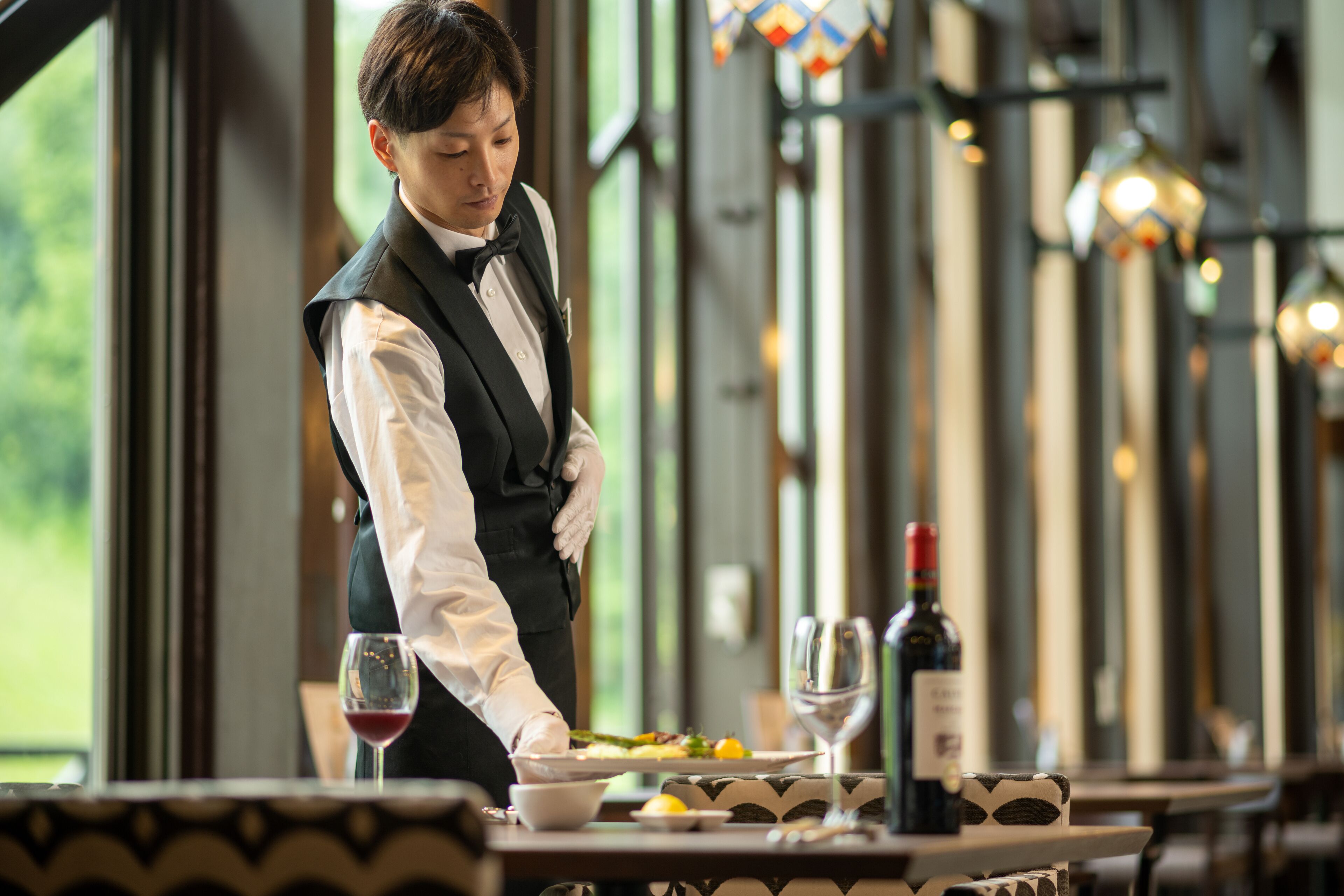 A waiter serving a meal at a restaurant with large windows and hanging lights.