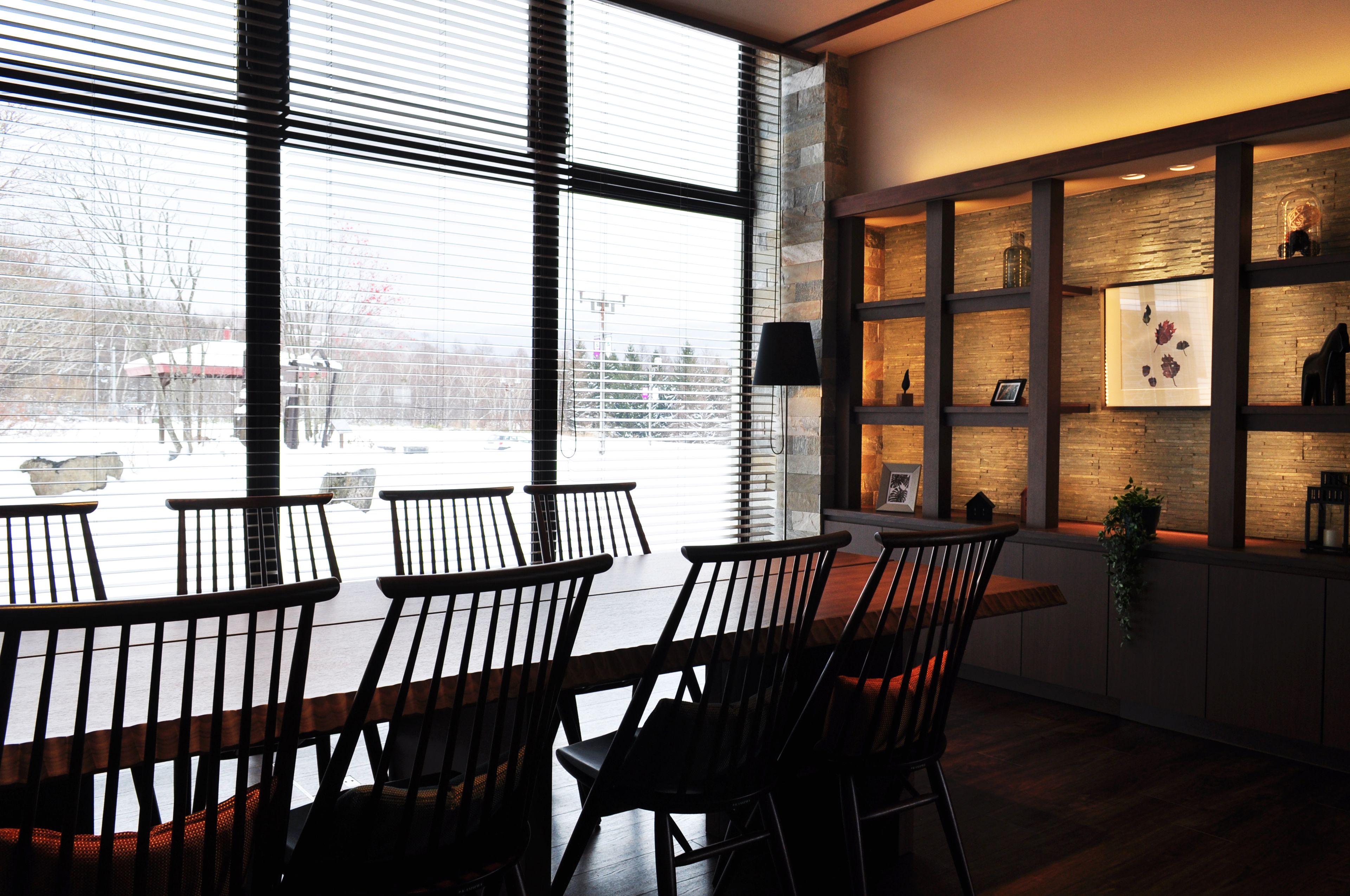 A dining area with a long table and chairs, a large window with blinds, and a wall with shelves displaying various items.
