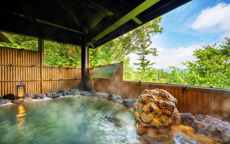 A hot spring bath area with a wooden deck, a stone rock formation, and a view of lush greenery.