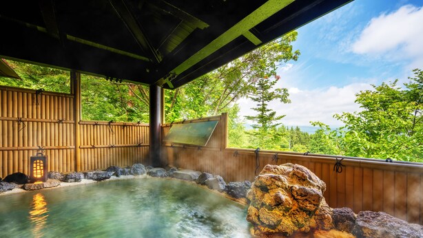 A hot spring bath area with a wooden deck, a stone rock formation, and a view of lush greenery.