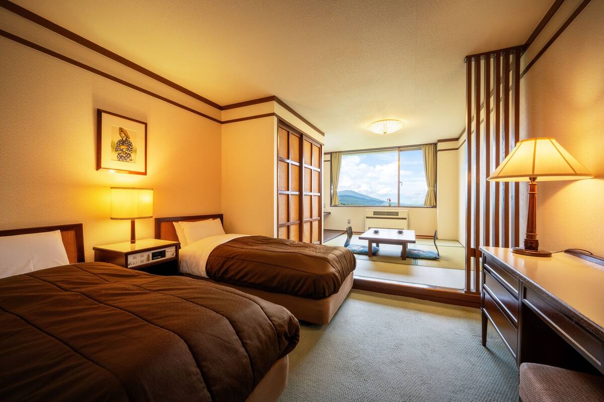A hotel room with two beds, a desk, a chair, a lamp, a window with a view, and a bedside table with a lamp.