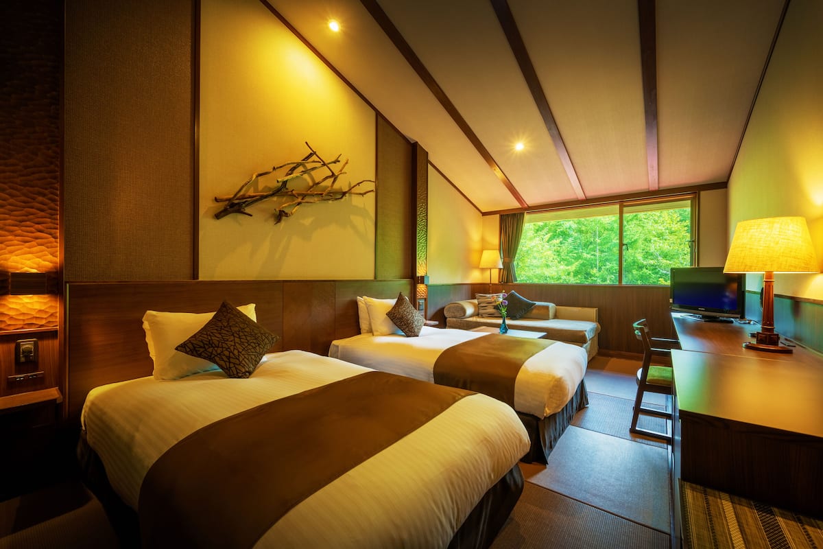A hotel room with two beds, a desk, a TV, and a window with a view of greenery.