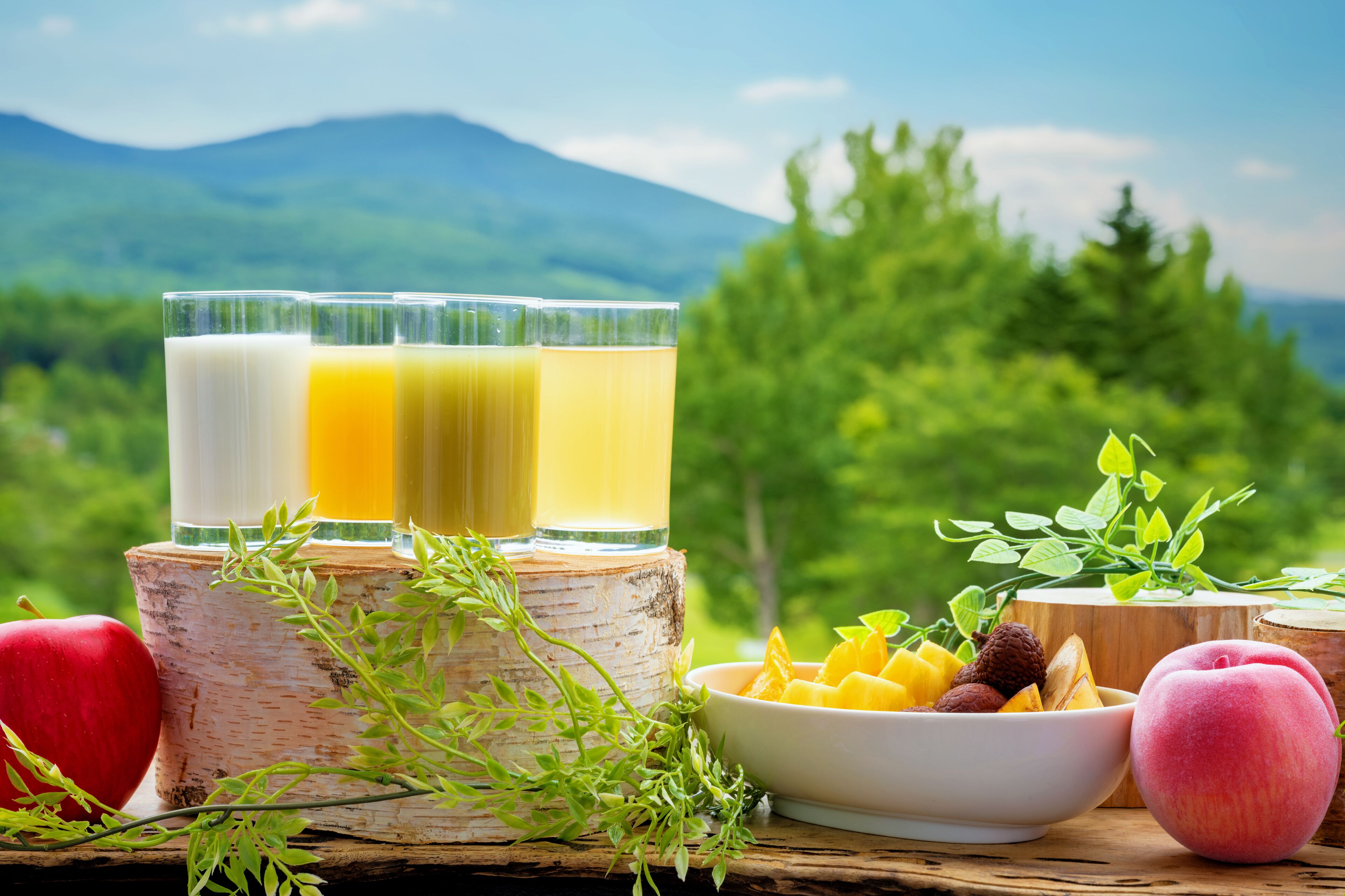 A wooden table with three glasses of juice, a bowl of fruit, and apples.