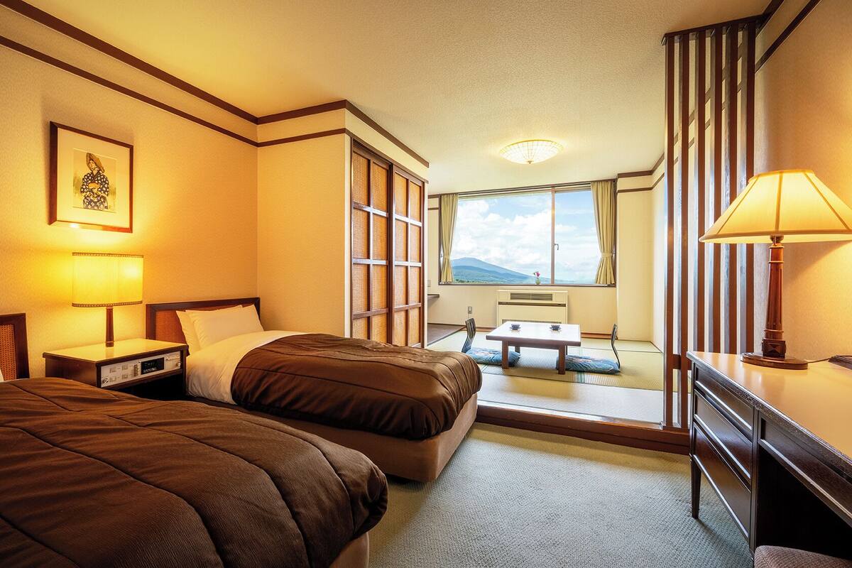 A hotel room with two beds, a desk, a chair, a window with a view, and a bedside lamp.