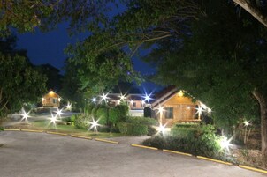 Vista nocturna de una cabaña con varias cabañas, rodeado de árboles e iluminado con luces exteriores.