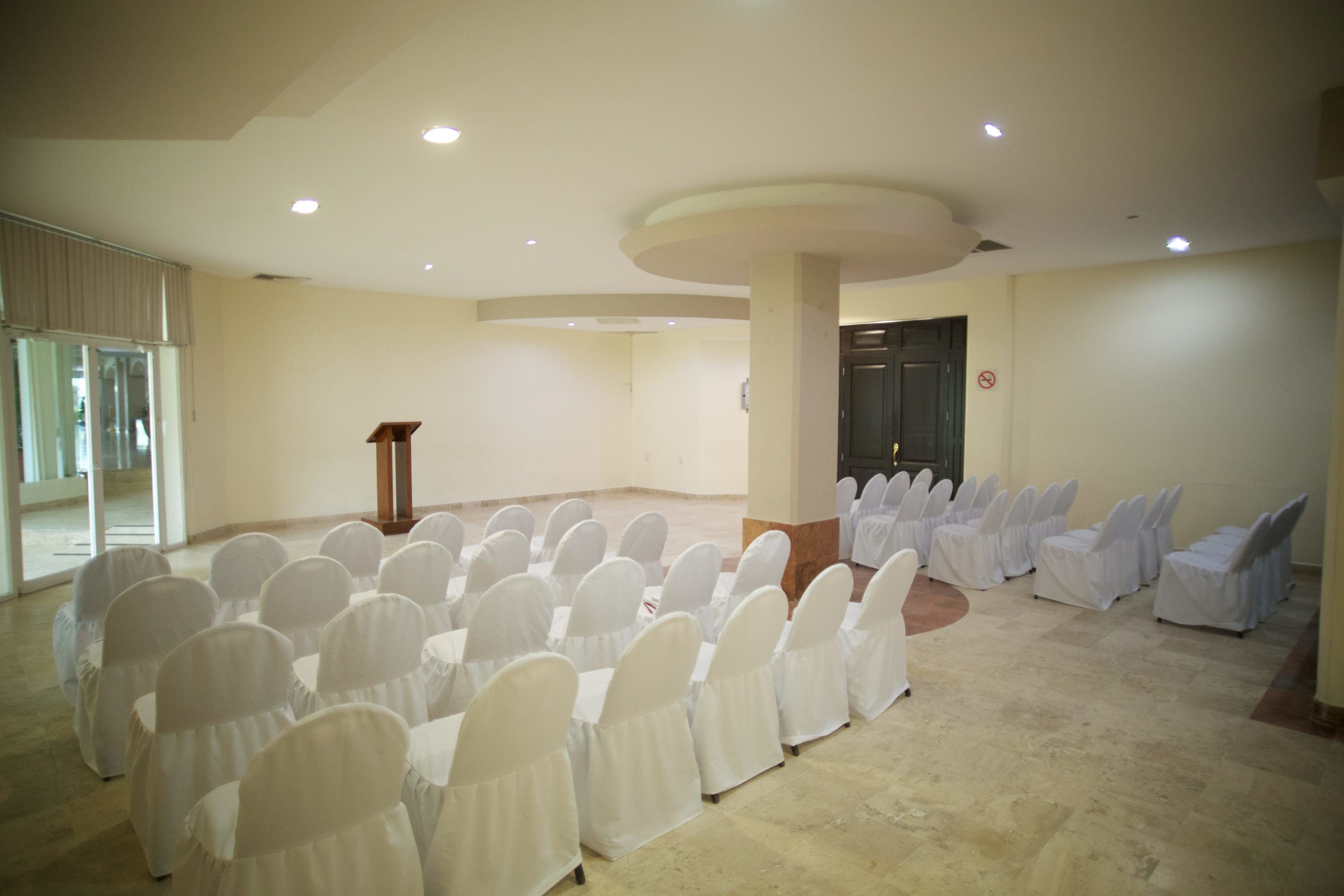 Un salón de conferencias con filas de sillas cubiertas con fundas blancas, un atril y una puerta con letrero de ‘prohibido el paso’.