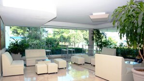 Un espacio interior moderno con sofás blancos y otomanas dispuestos alrededor de una mesa central. Amplias ventanas ofrecen vistas a un jardín exuberante y un coche azul en el exterior.