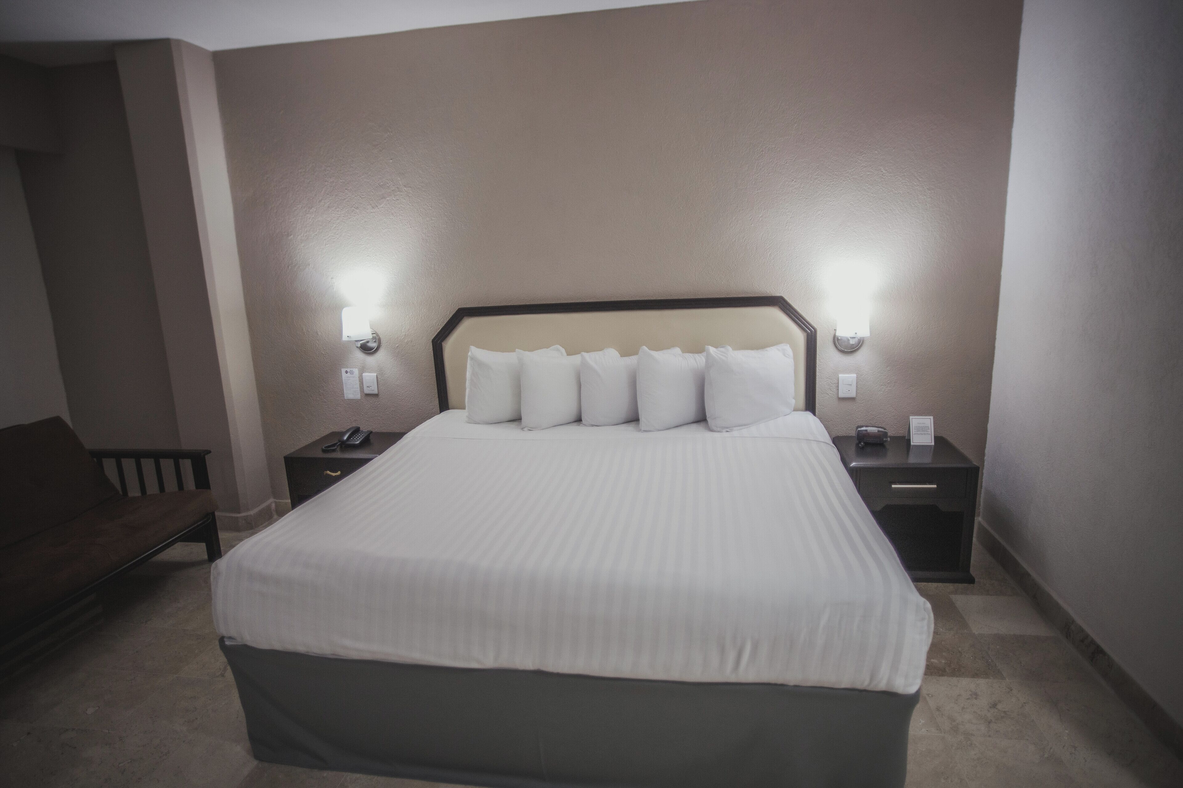 Quarto de hotel com uma cama grande, duas mesas de cabeceira e um banco.