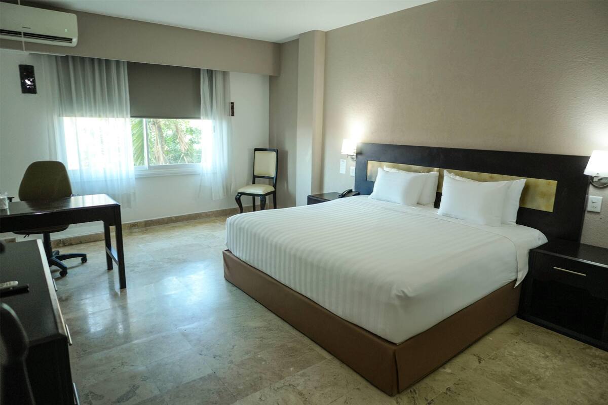 A hotel room with a large bed, a desk, a chair, and a window with curtains.