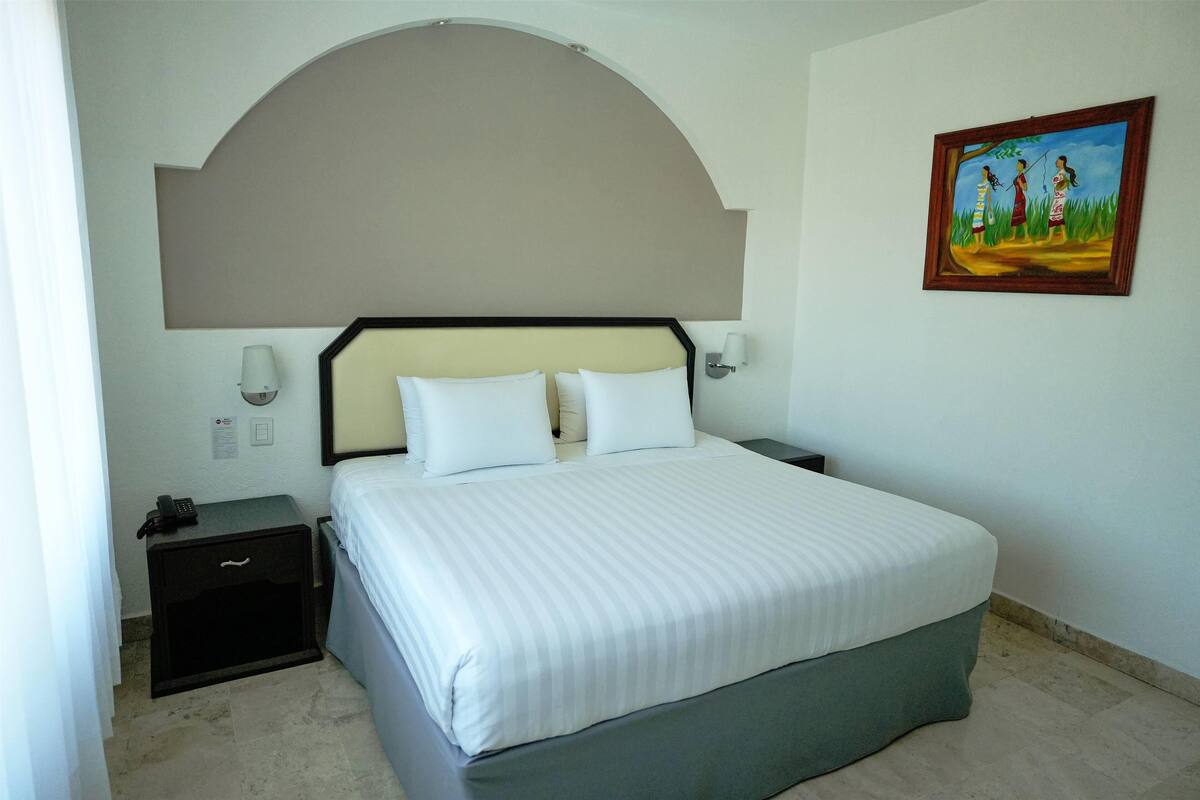A hotel room with a large bed, a nightstand, a lamp, and a painting on the wall.